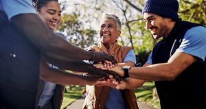 Smiling diverse team members stack hands in a collaborative gesture in a sunny park, conveying teamwork, unity, and shared enthusiasm.